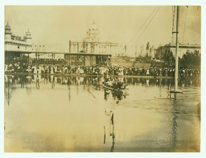 Horizontale foto van een zwemmer die een finishlijn passeert. Mannen in een boot kijken toe evenals menigten mensen aan de oever door Unknown